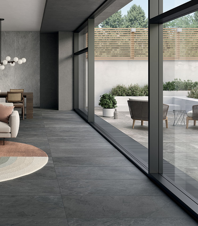 Zeitgemäßes Ambiente mit grauem Boden in Steinoptik, der Innen- und Außenbereich nahtlos verbindet, großen Glasflächen und einer schlicht gestalteten Terrasse.