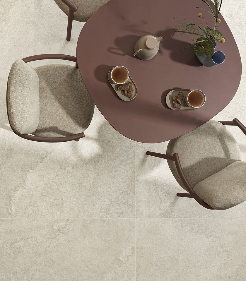 Runder terrakottafarbener Tisch mit Teeservice und beige gepolsterten Stühlen auf einem Boden aus der Kollektion Pulse, in Steinoptik im Farbton Shell.