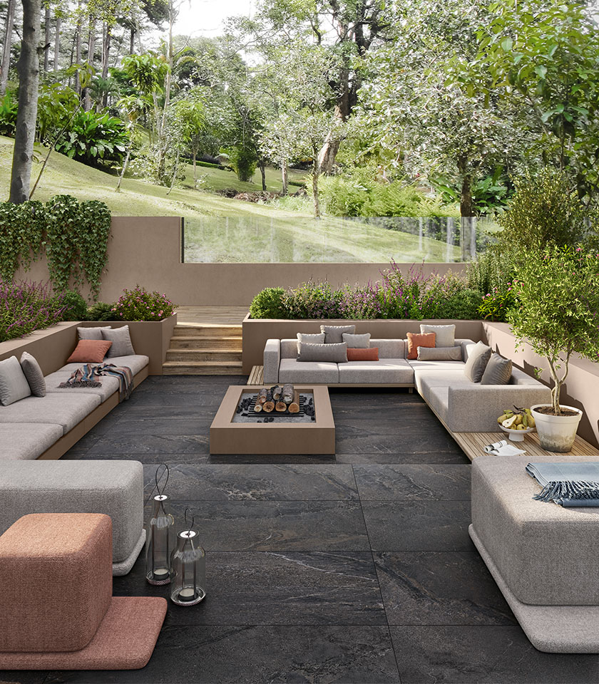 Terrasse im Grünen mit dunklem Steinoptik-Bodenbelag für den Außenbereich, großzügigen Lounge-Sitzmöbeln und integrierten Pflanzgefäßen für ein elegantes und einladendes Ambiente.