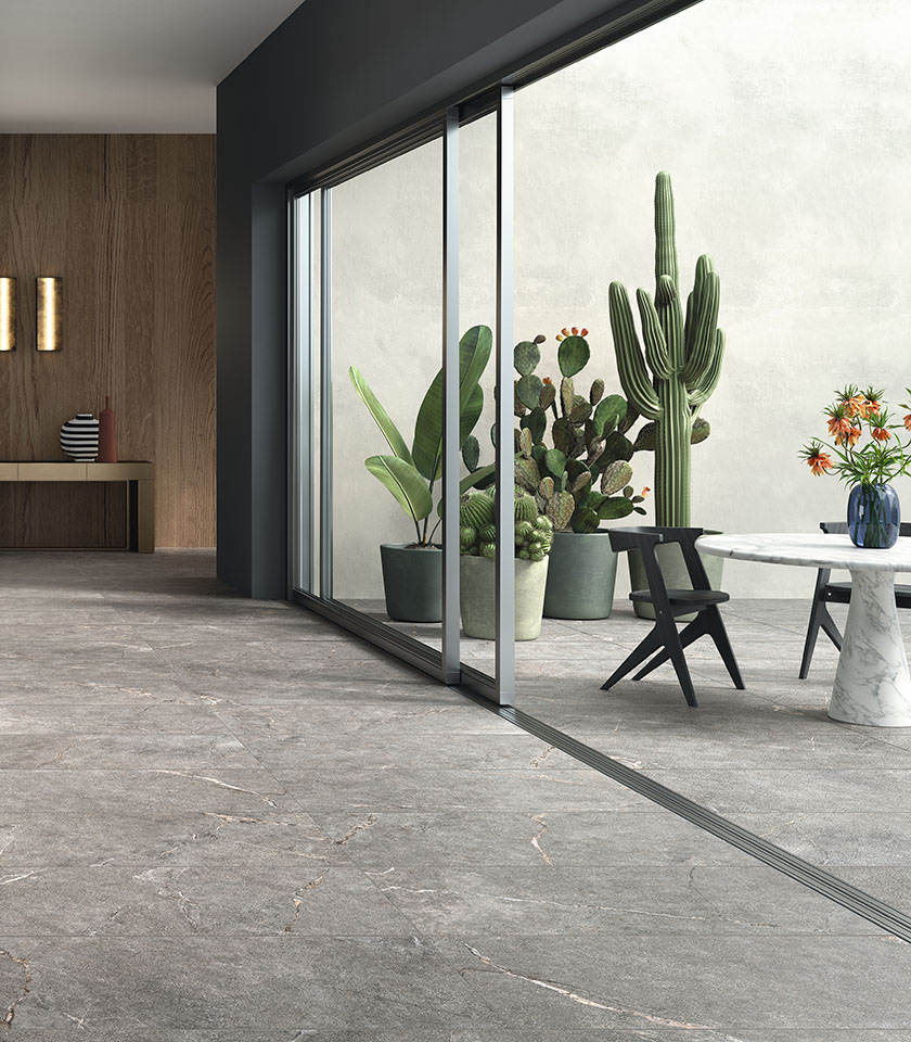 Zeitgemäßes Ambiente mit grauem Steinoptik-Bodenbelag, der Innen- und Außenbereich nahtlos verbindet, großen Glasflächen, puristischen Möbeln und dekorativen Pflanzen.